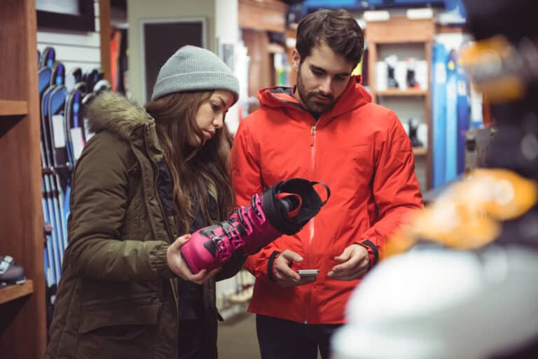 A woman in a beanie and winter coat holds a pink ski boot while a man in a red jacket checks its price tag inside a ski and winter sports shop. Skis and gear line the shelves around them.