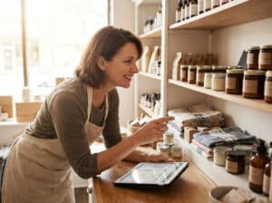 A smiling store manager in an apron leans on a wooden counter, pointing at stocked shelves while a tablet showing an inventory spreadsheet rests in front of her. The shelves hold amber jar candles, packaged goods, and folded linens in a warmly lit shop.